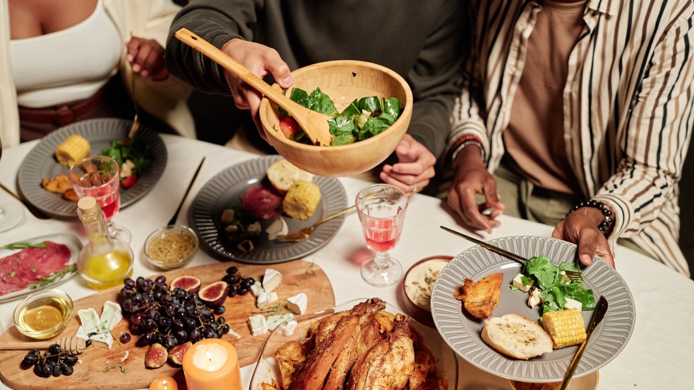 Masă împărțită între mai multe persoane, cu preparate simple și ingrediente proaspete, sugerând sustenabilitatea și dimensiunea socială a dietei mediteraneene.